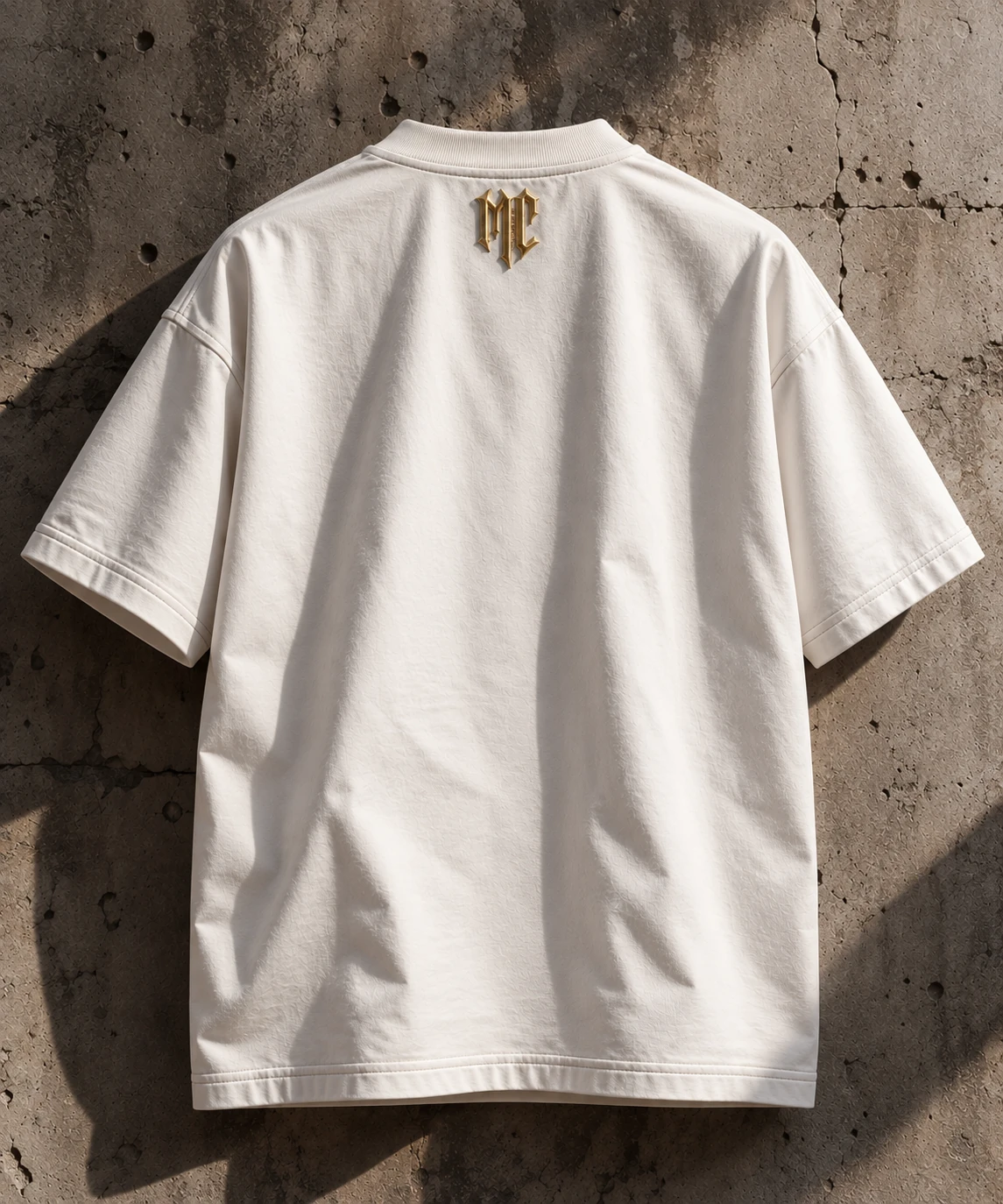 Back view of the white DIVINA T-shirt with the gold-toned M.C mark against a stone wall