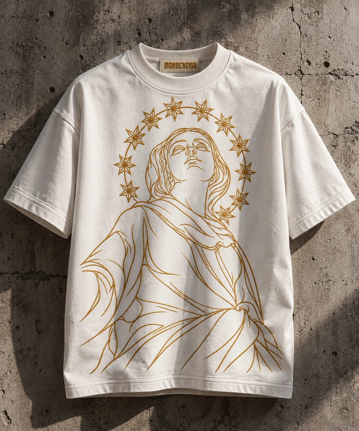 Front view of the white DIVINA T-shirt with a gold line-art figure and star halo against a stone wall